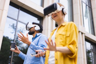 VR kulaklık takan iki kişi binanın yakınındaki artırılmış gerçekliği araştırıyor. Birinin elinde kahve fincanı ve telefonu var..