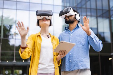 Bir adam ve bir kadın VR kulaklık takıyor ve dışarıda bir tabletle sanal gerçeklikle etkileşime geçiyorlar..