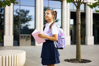 Güneşli bir günde okul binasının önünde at kuyruklu, sırt çantalı bir kız ve bir kitap duruyor..