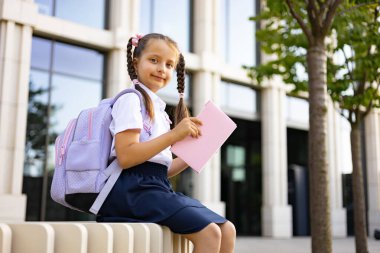 Örgülü örgülü saçları, sırt çantası ve pembe kitabı olan genç bir kız okul binasının önündeki duvarda oturuyor..
