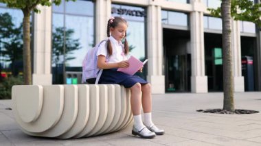Sırt çantalı genç bir kız okul binasının önündeki bankta oturup kitap okuyor..