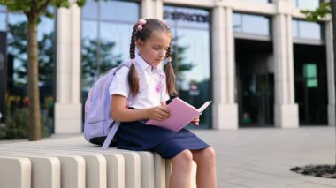Saçları örgülü ve sırt çantalı genç bir kız okulun önünde oturmuş pembe bir kitap okuyor..