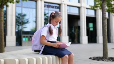 At kuyruklu ve sırt çantalı genç bir kız elinde kitapla okulun önünde oturuyor..
