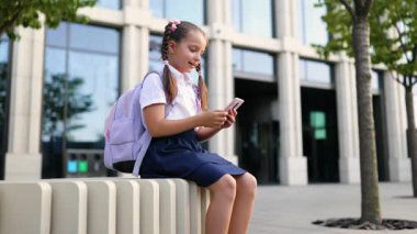 Sırt çantasıyla okul bahçesinde oturan ve telefonunu kullanan genç bir kız..