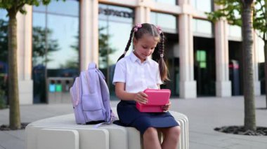 At kuyruklu beyaz bir kız okul binasının yanında sırt çantasıyla otururken abur cubur yiyor..