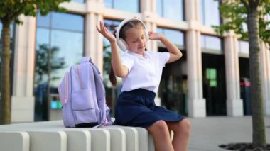 Okul bahçesinde, kulaklık takan, müzikten hoşlanan, arka planda bir okul binası olan beyaz bir kız öğrenci..