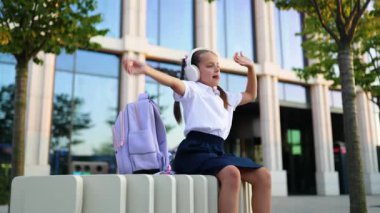 Beyaz bir kız öğrenci okul bahçesinde kulaklık takarak kollarını kaldırıyor..