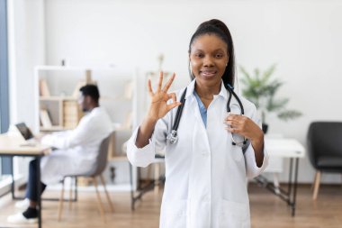 Sağlık ofisinde duran kendinden emin genç Afrikalı doktor. Steteskopu olan profesyonel bir kadın pozitif ifade sergiliyor. Arka planda sağlık çalışanı olan modern sağlık hizmetleri