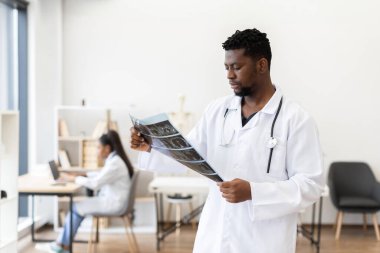 Beyaz önlüklü erkek doktor, muayenehanede röntgen taraması yaparken, kadın meslektaşı da iş yerinde meşgul. Profesyonel çevre, sağlık hizmetleri ekip çalışması ve teşhis sürecine örnek teşkil etmektedir..