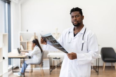 Erkek doktor, muayenehanede röntgen görüntüsünü inceliyor. Steteskoplu beyaz önlüklü genç bir erkek. Arka planda çalışan kadın meslektaşı, takım çalışması ve sağlık ortamını destekliyor.