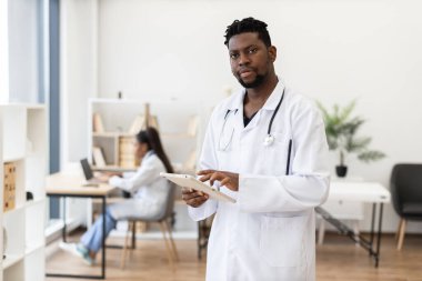Klinikte tablet kullanan erkek doktorun portresi ve arka planda çalışan bayan meslektaşı. Modern sağlık uygulamalarını, takım çalışmasını ve profesyonelliği yakalamak. Görüntü genç Afrikalı adamı içeriyor