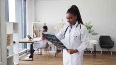 Genç bayan doktor röntgen görüntülerini tutuyor ve inceliyor. Profesyonel atmosferli çağdaş tıp ofisinde duruyor..