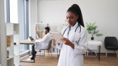 Steteskoplu beyaz önlüklü, kendinden emin Afrikalı kadın doktor klinik çalışma alanına başparmak işareti veriyor. Olumlu ifade profesyonelliği, güveni ve sağlık hizmetlerini ifade eder.