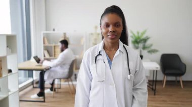 Genç Afrikalı kadın doktor portresi başparmakları havada duruyor. Sağlık hizmetleri profesyonelliğini ön plana çıkarıyor. Başka bir Afrikalı erkek doktoru da içeriyor