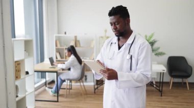 Beyaz önlüklü, elinde tablet olan bir erkek sağlık görevlisi. Geçmişinde kadın iş arkadaşı var. Tıbbi çalışma alanı belirleme, sağlık hizmetlerinde takım çalışması, teşhis teknolojisi, etkileşim.