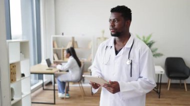 30 'lu yaşların başında erkek Afrikalı doktor ofiste tablet kullanıyor, iş arkadaşı yakınlarda çalışıyor, sağlık sigortası ayarında..