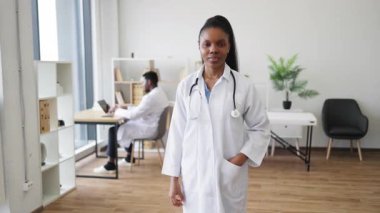 Sağlık ofisinde duran kendinden emin genç Afrikalı doktor. Steteskopu olan profesyonel bir kadın pozitif ifade sergiliyor. Arka planda sağlık çalışanı olan modern sağlık hizmetleri
