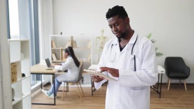 Beyaz önlüklü, kendine güvenen genç erkek tıp uzmanı, arka planda çalışan meslektaşı ile birlikte, klinik ortamda sağlık ekibi çalışmalarını ve modern sağlık teknolojisini ön plana çıkarıyor..