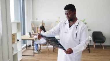 Beyaz önlüklü erkek doktor, muayenehanede röntgen taraması yaparken, kadın meslektaşı da iş yerinde meşgul. Profesyonel çevre, sağlık hizmetleri ekip çalışması ve teşhis sürecine örnek teşkil etmektedir..