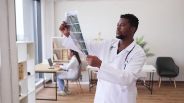 Modern ofis ortamında röntgen görüntülerini inceleyen erkek doktor, arka planda çalışan bayan meslektaşım. Radyoloji analizi ve teşhisinde görev alan profesyonel sağlık çalışanı.