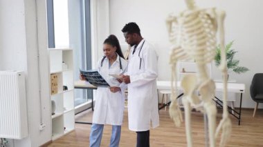 Modern klinikte erkek ve kadın doktorlar hastanın röntgenlerini inceliyor. Tıbbi araçlar ve iskelet modelinin yer aldığı profesyonel sağlık hizmetleri ortamı, sağlık uzmanlığını ve takım çalışmasını destekliyor