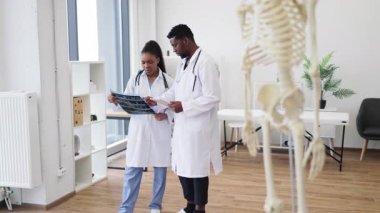 Laboratuvar önlüğü giyen erkek ve kadın doktorlar klinikte röntgen çekiyorlar. Ön planda görünen iskelet modeli, profesyonel çalışma alanı elementleriyle çevrili, tanılama iletiliyor