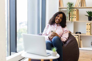 Armut koltukta rahat bir şekilde oturan genç bir kadın elinde akıllı telefonuyla doğal bir şekilde gülümsüyor. Arka planda büyük pencereli ve ev ofis kurulumlu parlak bir oda görünüyor..