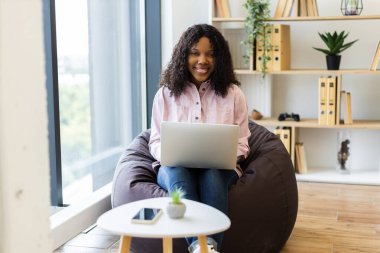 20 'li yaşlarda genç Afro-Amerikalı bir kadın sırt çantasına oturmuş gülümsüyor, pencere ve kitaplık kullanıyor, rahat ve rahat bir ev ortamı sağlıyor.