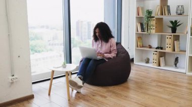 20 'li yaşlarda genç Afro-Amerikalı bir kadın sırt çantasına oturmuş gülümsüyor, pencere ve kitaplık kullanıyor, rahat ve rahat bir ev ortamı sağlıyor.