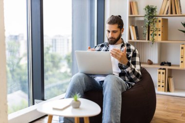 Genç, sakallı bir erkek. Ev ortamında dizüstü bilgisayarla çalışıyor. Büyük pencerenin yanında oturan fasulye torbası. Rahatlatıcı çalışma alanı kitap rafları, kapalı alan bitkisi, doğal ışık.