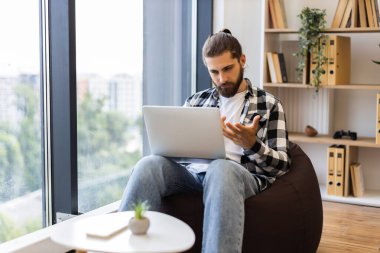 Genç, sakallı bir erkek. Ev ortamında dizüstü bilgisayarla çalışıyor. Büyük pencerenin yanında oturan fasulye torbası. Rahatlatıcı çalışma alanı kitap rafları, kapalı alan bitkisi, doğal ışık.