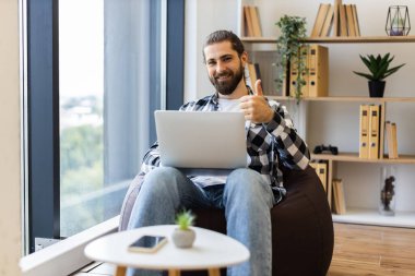 Sakallı, kareli gömlek giyen, dizüstü bilgisayarlı, parlak bir evde gülümseyen genç bir adam. Fasulye torbası, kapalı mekan bitkileri ve kitaplık da dahil olmak üzere rahat bir çalışma alanı. Erkek modeli yirmili yaşlarda gibi görünüyor..