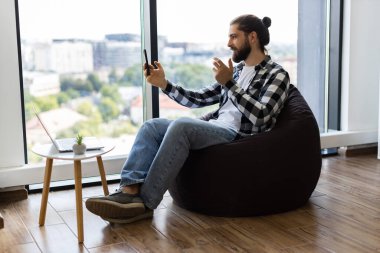 Ofiste puf minderinde oturan genç bir erkek iş günü akıllı telefondan sohbet ediyor. Parlak modern alandaki pencere görünümü uzak iletişim için gündelik, çoklu görev yaklaşımı tasvir eder.