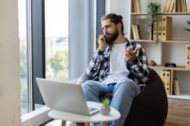 Geniş pencerenin yanında oturan sakallı, telefonda konuşan, gülümseyen genç bir erkek. Aydınlık modern oda, bitkiler, rahat görünüş. Dijital aygıtlarla ev konseptinden uzak çalışma.