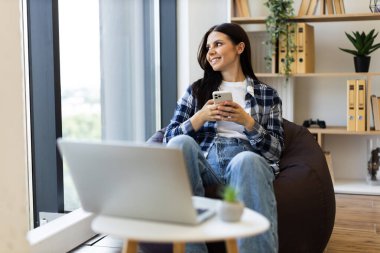 Kafkasyalı genç bir kadın, şık, parlak oturma odasında dekorlu akıllı telefon kullanırken gülümsüyor. Sıradan kıyafetler giymek, pencerenin yanındaki rahat koltuğun keyfini çıkarmak, neşeli duygular sergilemek..