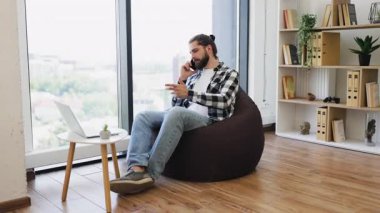 Çağdaş odada fasulye çuvalının üzerinde oturan genç adam telefonla konuşuyor, uzaktan çalışıyor..