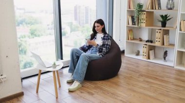 Sıradan kıyafetler giyen genç bir yetişkin pencerenin yanındaki akıllı telefonu kullanarak fasulye çantasının üzerinde oturuyor. İç kısımda kitaplar ve bitkilerle dolu raflar, ahşap zemin ve parlak gün ışığı yer alıyor..