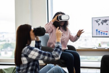 Focused image of women in their twenties exploring virtual reality with headsets indoors. Enhanced tech-oriented environment with charts visible on screen