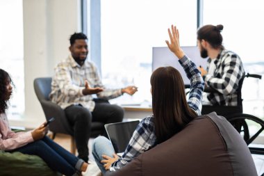 Young adults discussing concepts in relaxed office setting, woman raising hand to ask question. Individuals of diverse racial backgrounds, including person using wheelchair highlighting inclusivity.