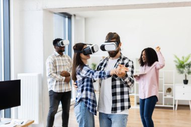 Diverse group of young professionals enjoying activities with virtual reality glasses inside office setting. Two dancing while others laugh, demonstrating happiness and creativity in technology.