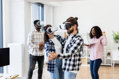 Diverse group of young professionals enjoying activities with virtual reality glasses inside office setting. Two dancing while others laugh, demonstrating happiness and creativity in technology.