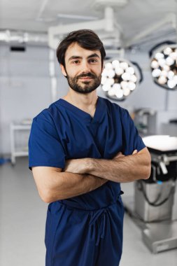 A surgeon stands with arms crossed in an operating room, looking at the camera.
