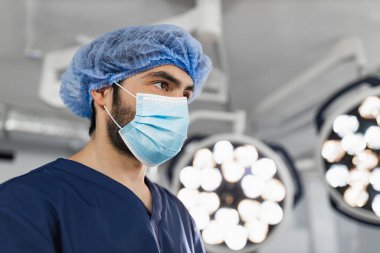 A surgeon wearing a surgical mask and cap in an operating room, illuminated by bright lights.