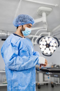 A surgeon in a surgical mask and scrubs prepares for an operation in a modern operating room.