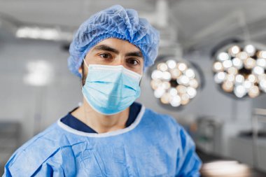 A surgeon wearing a surgical mask and cap stands in an operating room, illuminated by bright lights.