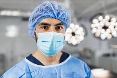A surgeon wearing a surgical mask and cap in an operating room, looking directly at the camera.