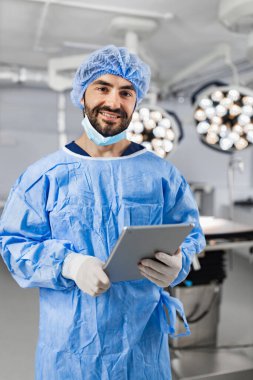 A smiling surgeon in scrubs holds a tablet in an operating room, ready for a procedure.