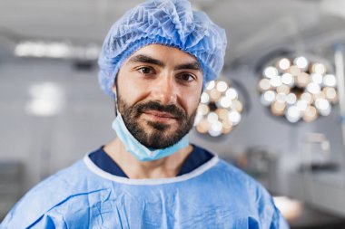 A surgeon, wearing a surgical cap and mask, stands in an operating room, looking at the camera.