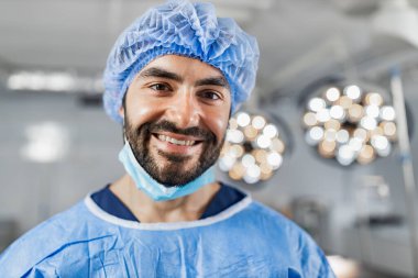A cheerful surgeon smiles directly at the camera in a bright operating room, wearing surgical attire.
