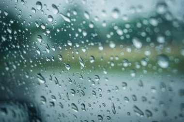 Fırtınalı bir günde yağmur damlalarının şehir manzarası ön cama düşer. Bulanık binalar trafik ışığı arka planında yükselir..